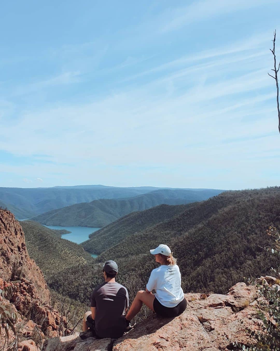 Two people sitting on a mountain lookout overlooking forested valleys and a lake.